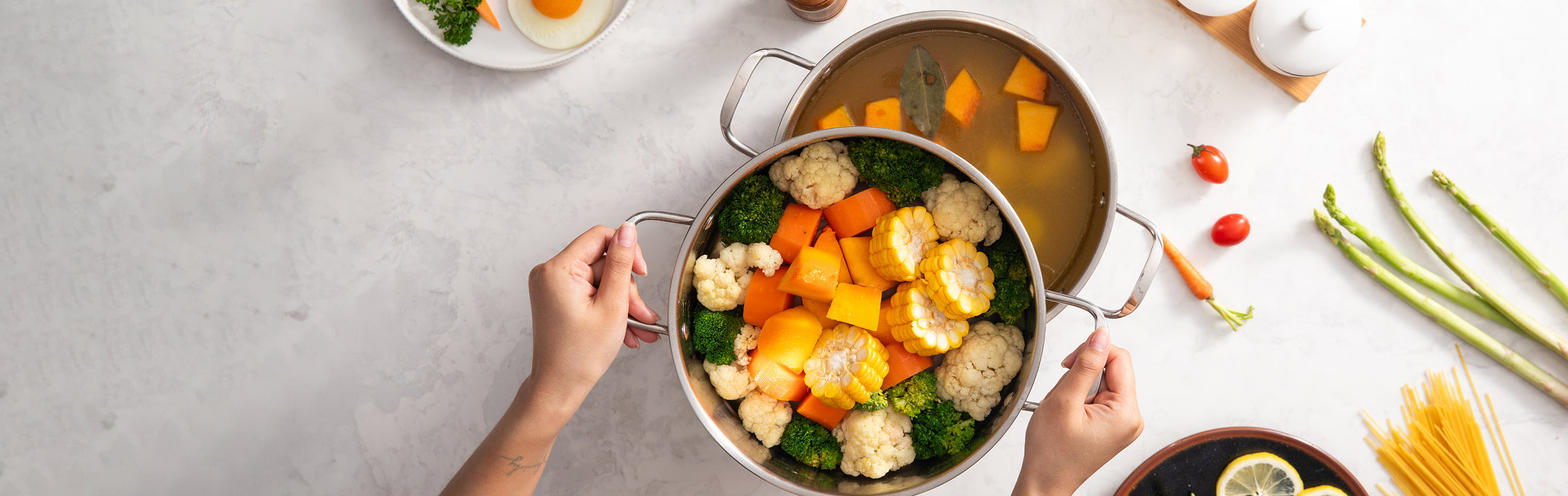Person holding a pot of vegetable soup with various vegetables on a light surface.